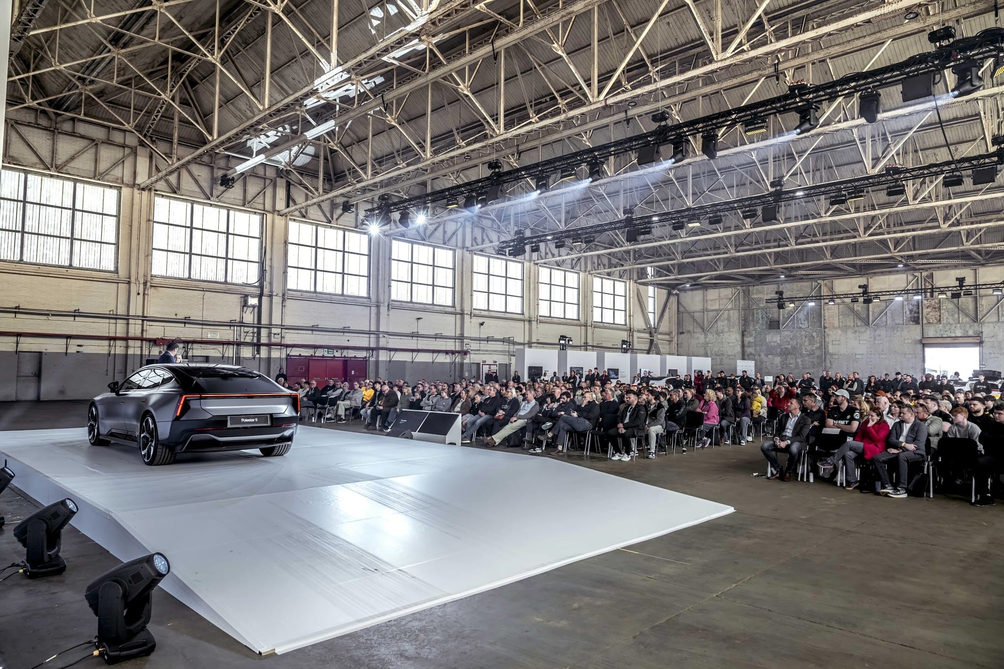 Polestar 5 rear three-quarter on the white stage with audience and hangar truss, Polestar Festival, September 2025.